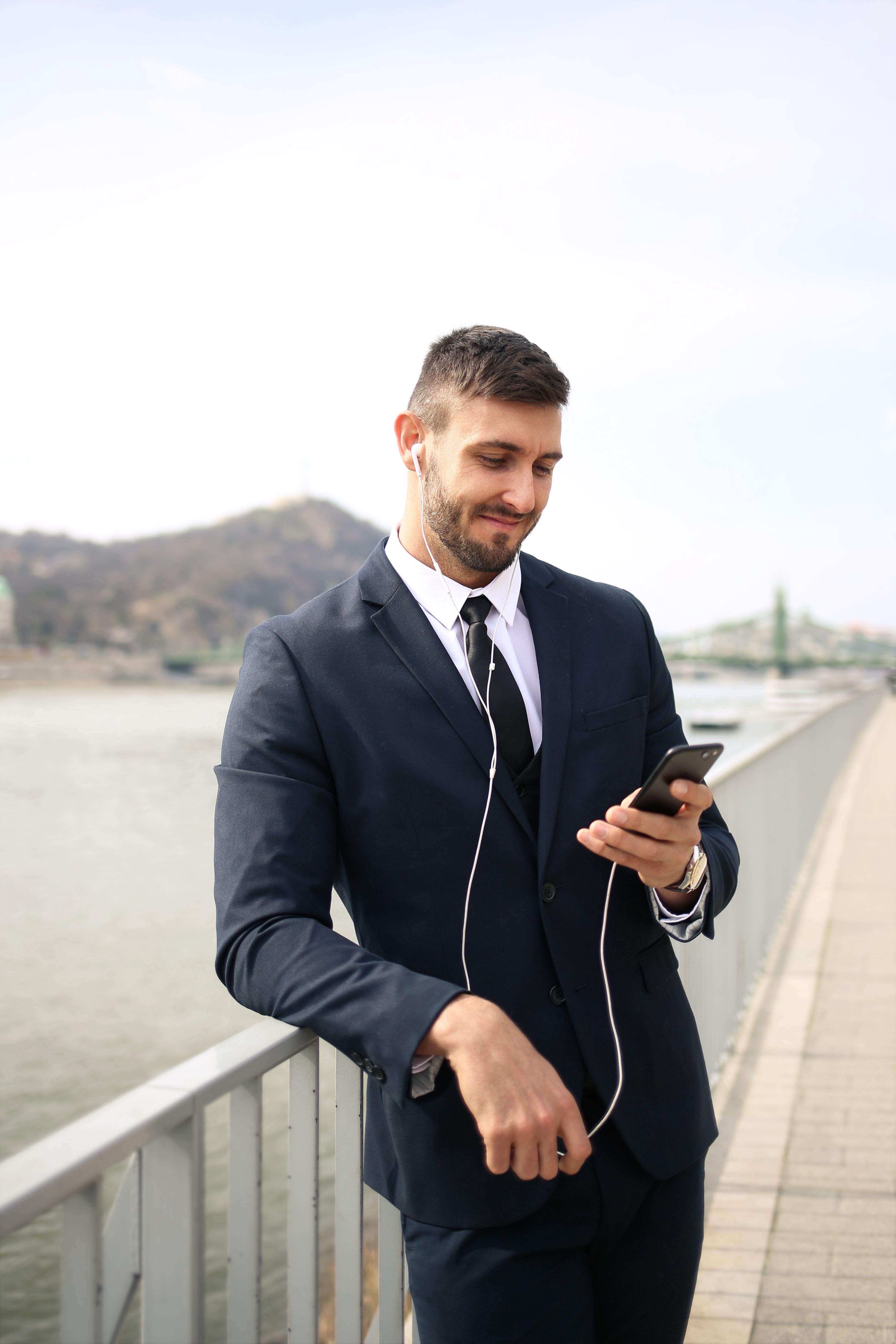 Man in a dark suit and earbuds smiling at his phone on a waterfront walkway